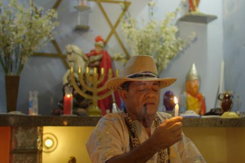Homem com chapéu de palha e colares fuma cachimbo e aparece em frente a um altar religioso, segurando uma vela acesa com expressão de concentração e respeito. O altar é decorado com flores, imagens de santos e entidades, velas coloridas e um candelabro dourado, compondo um ambiente espiritual e ritualístico.
