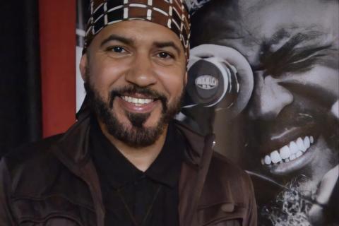 O pedagogo, curador, cineasta, roteirista e produtor Alek Lean sorri para a câmera, usando camisa preta, jaqueta marrom e um turbante estampado em marrom e branco. De barba bem aparada e expressão acolhedora, ele posa diante de um painel em preto e branco que mostra, em detalhe, o rosto de um homem sorridente, criando um cenário de forte atmosfera artística.