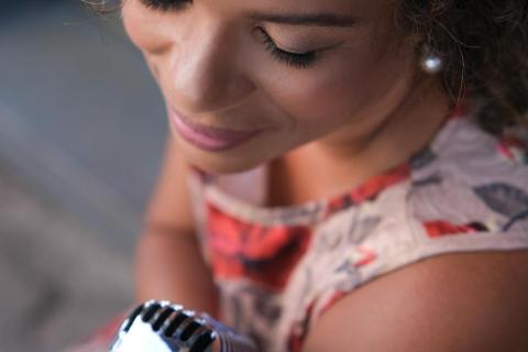 A imagem mostra a cantora Debora Ondina vista de cima e de lado, com expressão serena enquanto segura um microfone vintage prateado. Morena clara, de cabelos cacheados curtos e usando um vestido estampado em tons suaves, ela transmite delicadeza e concentração, como se estivesse prestes a cantar.