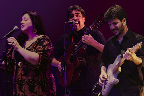 A imagem mostra os músicos Luiza Borges (voz), Thiago Thiago de Mello (voz e violão) e Diogo Sili (guitarras e arranjos) em apresentação. Luiza canta com expressão serena, Thiago interpreta com intensidade ao violão e Diogo toca guitarra com concentração. O fundo em tons de roxo e vermelho valoriza a cena, destacando a energia e harmonia do trio no palco.