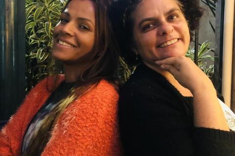 As escritoras Luciana Belém e Ednéia Rodrigues posam sorridentes, encostadas uma na outra, diante de um fundo com plantas. Luciana, à esquerda, veste um casaco alaranjado e tem cabelos longos; Ednéia, à direita, usa um casaco preto e apoia o queixo na mão, com expressão acolhedora. A imagem transmite leveza e cumplicidade entre as autoras.