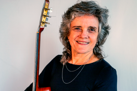 A violonista Vera de Andrade, de cabelos grisalhos e ondulados, sorri suavemente enquanto segura o violão apoiado sobre o ombro. Ela veste uma blusa escura e um colar delicado de duas voltas. O fundo neutro realça sua expressão serena e confiante, refletindo sua conexão com o instrumento e sua trajetória musical.