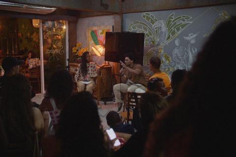A imagem mostra um espaço cultural intimista, com pessoas sentadas assistindo a uma roda de conversa ou apresentação. À frente, duas pessoas estão no palco: uma mulher de cabelos longos e um homem falando ao microfone. O ambiente é decorado com murais artísticos na parede, iluminação aconchegante e plantas, criando uma atmosfera acolhedora e descontraída.