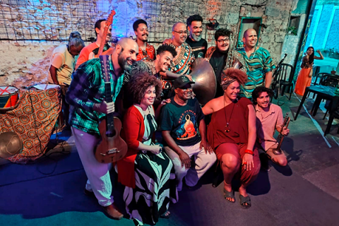 Integrantes da Orquestra Balogun posam juntos em um ambiente rústico, com paredes de pedra e iluminação colorida em tons de azul e vermelho. O grupo reúne músicos e artistas com instrumentos como violão, atabaque e outros tambores, formando uma composição vibrante. Todos sorriem e se aproximam para a foto, transmitindo união, celebração e a força da musicalidade afro-brasileira que marca o trabalho da orquestra.