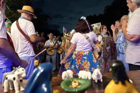 Integrantes de Folia de Reis do Sertão Carioca se reúnem ao ar livre à noite, vestidos com roupas coloridas, chapéus e instrumentos musicais como violas e pandeiros. Em primeiro plano, um presépio artesanal com figuras de barro representa o nascimento de Jesus. Ao fundo, o grupo canta e toca em celebração tradicional, marcada pela devoção, alegria e forte expressão da cultura popular.