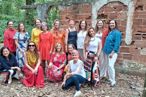 Integrantes do Acorde Vocal, grupo vocal feminino, posam juntas ao ar livre diante de uma antiga parede de tijolos aparentes. As mulheres vestem roupas coloridas e variadas, algumas em pé e outras agachadas, sorrindo para a câmera. O cenário natural, com folhas e vegetação ao redor, transmite acolhimento, união e a força coletiva do grupo.