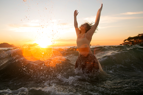 Mulher está no mar, com a água chegando à cintura, de braços erguidos em gesto de celebração. Ela usa blusa clara e saia estampada, enquanto ondas se quebram ao redor. Ao fundo, o sol se põe no horizonte, criando luz dourada e reflexos na água, transmitindo sensação de liberdade, alegria e conexão com a natureza.