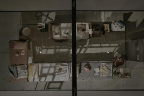 Imagem vista de cima de uma cela coletiva, mostrada como cena do documentário Torre das Donzelas: Memórias e Resistências de Presas Políticas da Ditadura Militar. O espaço é dividido por grades e sombras projetadas, com colchões finos no chão, cobertores dobrados e poucos objetos pessoais espalhados, como livros, roupas e recipientes. A composição rígida e simétrica transmite sensação de confinamento, vigilância e precariedade, reforçando o contexto de repressão e resistência vivido pelas presas políticas.