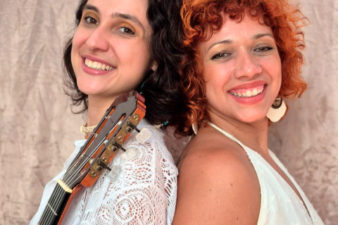 Fotografia de duas mulheres sorrindo, apoiadas costas com costas, diante de um fundo neutro. À esquerda está a violonista Maria Luísa Tonácio, de pele clara e cabelo escuro ondulado, usando blusa branca rendada; o braço de um violão aparece apoiado em seu ombro. À direita está a cantora Deya Mota, de pele clara, cabelos curtos e cacheados em tom ruivo, usando brinco claro e blusa clara sem mangas. Ambas olham para a câmera com expressão alegre.