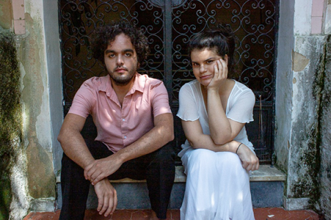 Fotografia de retrato do Duo Bevilacqua Assumpção (violoncelo/piano). A dupla está sentados lado a lado em um degrau, diante de uma porta com grade de ferro trabalhado. À esquerda, o homem de pele clara com cabelos cacheados e barba, veste camisa rosa e calça escura, olhando para a câmera com expressão séria. À direita, a mulher de pele clara com cabelos presos, veste roupa clara e apoia o rosto na mão, sorrindo suavemente. O ambiente tem paredes com marcas do tempo, sugerindo cenário externo ou histórico.