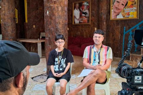 Cena do documentário Dos Armários para as Ruas mostra duas pessoas sentadas lado a lado para uma entrevista. À esquerda, uma criança de óculos, camiseta preta e colar; à direita, um adulto com camisa colorida listrada, short e chinelos, segurando um papel. Em primeiro plano, parte de um cinegrafista e uma câmera profissional indicam a gravação. O ambiente interno tem paredes de pedra, colunas e quadros nas paredes, com clima íntimo e documental.