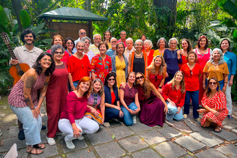 Fotografia de grupo do Coral da Tarde em Canto Plural ao ar livre. Cerca de trinta integrantes estão reunidos em um jardim, entre árvores e plantas, organizados em fileiras — alguns em pé e outros agachados à frente. O grupo é diverso em idade e aparência, vestindo roupas coloridas, com predominância de tons quentes. Todos sorriem e olham para a câmera, transmitindo um clima de alegria e convivência.