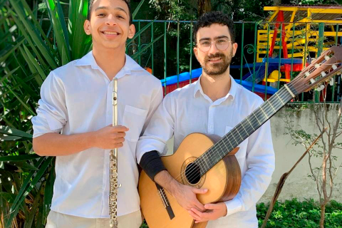 Fotografia de retrato do Duo Sereno, formado pelo flautista Vinícius Marinho e o violonista Iuri Chicharo. Os dois estão lado a lado ao ar livre, sorrindo para a câmera. À esquerda, Vinícius, homem de pele clara, usa camisa branca e segura uma flauta transversal. À direita, Iuri, homem de pele clara com barba e óculos, também veste camisa branca e segura um violão. Ao fundo há vegetação e uma área com brinquedos coloridos, sugerindo um espaço externo ensolarado.