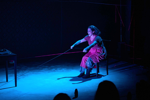 Cena da peça Filipa mostra uma mulher sentada em uma cadeira, em palco escuro iluminado por luz azul. Ela veste vestido vermelho e puxa com força um fio ou corda esticada à sua frente, com expressão intensa e concentrada. O cenário é minimalista, com poucos objetos, e linhas finas coloridas atravessam o espaço, criando tensão visual e reforçando o clima dramático da cena.