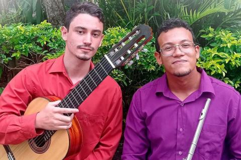 Retrato dos dois músicos do Duo Prevatto & Do Carmo ao ar livre, diante de folhagens. À esquerda, Tacio Prevatto segura um violão clássico, veste camisa vermelha e olha para a câmera com expressão tranquila. À direita, Erick do Carmo segura uma flauta transversal, usa camisa roxa e óculos, sorrindo levemente. A cena tem luz natural e clima informal, destacando a dupla e seus instrumentos.