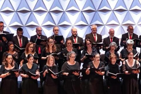 Apresentação de coral em palco mostra grupo numeroso de cantores organizados em fileiras. Todos vestem roupas formais em tons escuros, com alguns homens usando gravatas vermelhas. Cada integrante segura uma partitura e canta com expressão concentrada. Ao fundo, um painel geométrico branco cria textura visual. A iluminação é clara e uniforme, destacando o conjunto e a harmonia da apresentação.