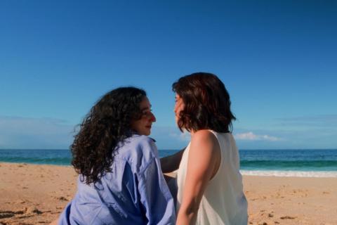 Cena do filme Azul mostra duas mulheres sentadas na areia de uma praia, próximas ao mar. Elas estão de perfil, voltadas uma para a outra, com expressões suaves e íntimas. Uma veste camisa azul e tem cabelos longos e cacheados; a outra usa blusa clara e tem cabelo curto. Ao fundo, o mar azul e o céu aberto criam um clima tranquilo e contemplativo.