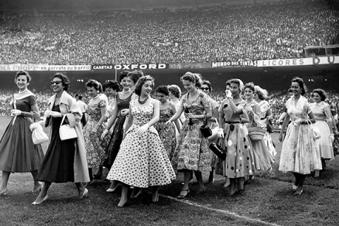 Imagem em preto e branco do documentário “Um filme à moda Bangu”. Um grupo de mulheres com vestidos rodados e estampados típicos dos anos 1950 caminha pelo gramado de um estádio lotado. Algumas sorriem e conversam entre si, enquanto a multidão aparece ao fundo nas arquibancadas. A cena remete ao universo da moda, do cotidiano e da cultura popular brasileira da época.