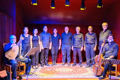 Integrantes do Coro Madrigal da ACC posam em fotografia. O grupo está alinhado diante de uma cortina vermelha, vestindo trajes pretos. Nas extremidades, dois músicos seguram instrumentos de cordas antigos, um alaúde e uma viola da gamba. A iluminação cênica em tons quentes destaca os participantes e o tapete ornamentado que compõe o cenário, criando atmosfera de concerto de música vocal e de câmara.