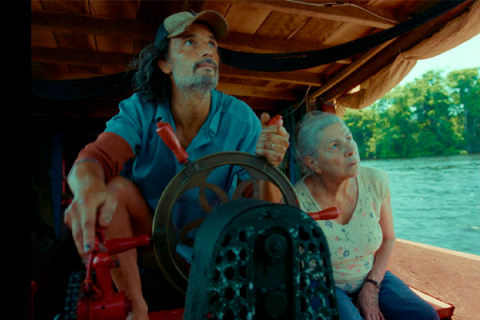 Cena do filme “O Último Azul”, com os atores Rodrigo Santoro e Denise Weinberg. Em uma embarcação sobre um rio, Rodrigo Santoro, de boné e camisa azul, segura o leme enquanto observa algo à frente. Ao seu lado, Denise Weinberg acompanha a mesma direção com expressão atenta. Ao fundo, aparecem a água e a vegetação ribeirinha, reforçando a atmosfera de viagem pelos rios da Amazônia.