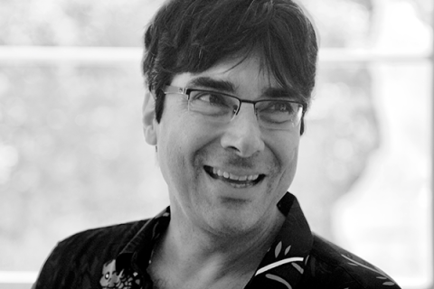 Retrato em preto e branco do poeta Alberto Pucheu. Ele aparece em primeiro plano, usando óculos e camisa escura estampada, com um sorriso espontâneo enquanto olha para fora do enquadramento. O fundo desfocado e a iluminação suave destacam sua expressão acolhedora e a atmosfera descontraída da fotografia.