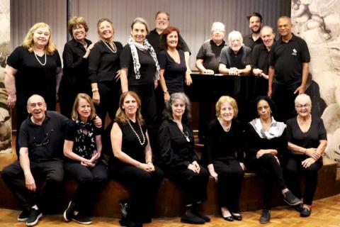 Integrantes do Coro Tu Voz, Mi Voz, da Associação de Canto Coral, posam reunidos em um palco. O grupo é formado por homens e mulheres de diferentes idades, distribuídos entre pessoas sentadas à frente e outras em pé ao fundo, todas vestindo roupas predominantemente pretas. Ao centro, um piano reúne parte dos integrantes, enquanto o piso de madeira e as cortinas ao fundo compõem o ambiente da apresentação.