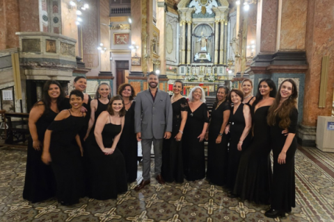 Integrantes do Coro Lírico Feminino (ACC) posam ao lado do maestro Cláudio Ávila no interior de uma igreja. O maestro, de terno cinza, está ao centro, cercado pelas cantoras, que vestem longos vestidos pretos. Ao fundo, o altar ornamentado, colunas de mármore e lustres iluminados destacam a arquitetura do templo.