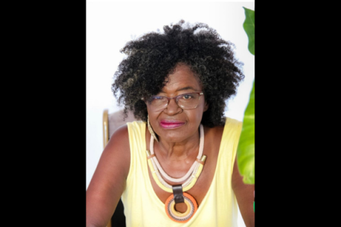 Retrato da escritora e poeta Miriam Alves. Ela olha diretamente para a câmera, com expressão serena e leve sorriso. Tem pele negra, cabelos crespos volumosos com fios grisalhos, usa óculos de armação discreta, blusa amarela sem mangas e um colar de grandes argolas em tons de bege, marrom e laranja. Ao fundo, há uma parede clara e, à direita da imagem, parte de uma folha verde aparece em primeiro plano.