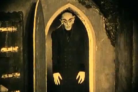 Cena do filme Nosferatu. Um homem de aparência cadavérica, com cabeça raspada, orelhas pontiagudas e dedos longos, surge parado à entrada de uma porta em arco, envolto por sombras. Vestido com roupas escuras, mantém a cabeça levemente inclinada e os braços estendidos ao lado do corpo. A iluminação em tons amarelados e o contraste intenso entre luz e escuridão reforçam a atmosfera sombria da cena.