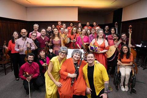 Integrantes da Orquestra Barroca da UNIRIO posam reunidos em uma sala de concertos. O grupo é formado por músicos de diferentes idades, distribuídos em várias fileiras, alguns segurando instrumentos de cordas, sopro e dedilhados. Em primeiro plano, três integrantes vestindo roupas em tons de amarelo e laranja ocupam o centro da composição, enquanto, ao fundo, os demais sorriem para a câmera. A imagem destaca a diversidade do conjunto e a atmosfera de celebração entre os músicos.