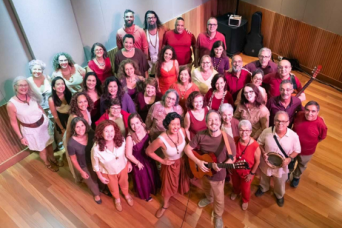 Integrantes do coro Ordizão posam para uma fotografia em grupo em uma sala de ensaio com piso de madeira. Cerca de quarenta participantes, de diferentes idades, sorriem para a câmera, distribuídos em várias fileiras e fotografados de um ângulo alto. As roupas em tons de vermelho, vinho, rosa, marrom e bege criam uma composição harmoniosa e acolhedora. Em primeiro plano, um integrante segura um violão, enquanto outro segura um pandeiro. Ao fundo, encostados na parede, aparecem um violão e um amplificador.
