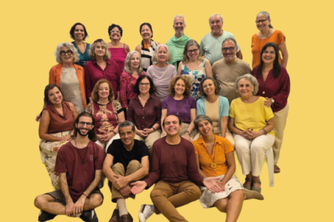 O Coral Pedra da Urca reúne cerca de vinte e cinco integrantes em um retrato coletivo sobre fundo amarelo-claro. Os participantes, distribuídos em quatro fileiras, sorriem para a câmera e vestem roupas em cores variadas, predominando tons quentes e coloridos. Alguns estão em pé, outros sentados e três aparecem acomodados no chão à frente do grupo, compondo uma fotografia descontraída.