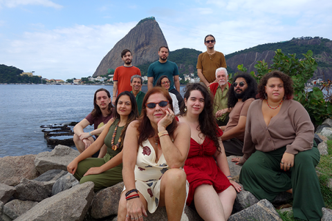Integrantes do Coral Altivoz posam reunidos sobre pedras à beira da água, tendo ao fundo o Pão de Açúcar e a paisagem da Baía de Guanabara. O grupo é formado por pessoas de diferentes idades, distribuídas em vários níveis, algumas sentadas e outras em pé. Vestindo roupas em tons terrosos, verdes, vermelhos e bege, os integrantes sorriem ou mantêm expressões serenas, compondo um retrato coletivo em ambiente ao ar livre.
