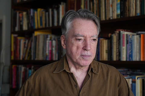 O poeta Braulio Tavares é retratado diante de uma estante repleta de livros. Com cabelos grisalhos penteados para trás e camisa marrom, mantém o olhar voltado levemente para baixo, com expressão serena e reflexiva. O fundo desfocado destaca as prateleiras repletas de livros, reforçando o ambiente de leitura e criação literária.