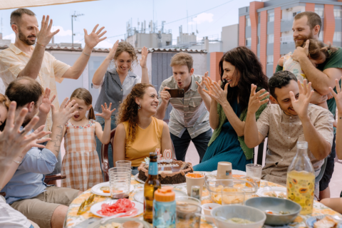 Still de um dos filmes da mostra de cinema espanhol. Um grupo de adultos e uma criança está reunido ao redor de uma mesa durante uma comemoração. Ao centro, uma mulher sorri diante de um bolo com velas que formam o número 40, enquanto as pessoas ao redor levantam as mãos em gestos animados. A mesa está repleta de pratos, copos e bebidas, e a cena acontece em uma área externa cercada por edifícios.