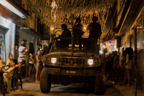Still de filme. À noite, um veículo militar com os faróis acesos ocupa o centro de uma rua estreita, com homens fardados na parte superior. Moradores observam a movimentação nas calçadas e diante das casas. Acima da rua, numerosas fitas ou bandeirolas de festa decoram o espaço, iluminado por luzes em tons amarelados.