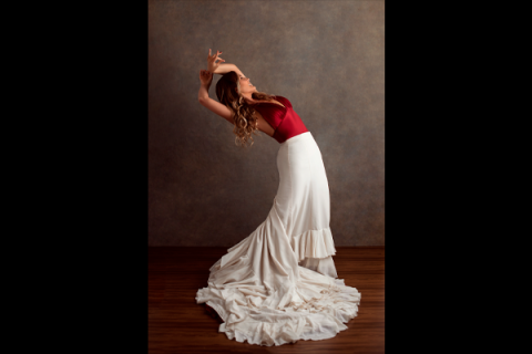 Retrato da bailarina Eliane Carvalho em pose de dança. Ela inclina o corpo para trás, com o rosto voltado para cima e os braços elevados e curvados sobre a cabeça. Veste blusa vermelha e uma longa saia branca com babados, cuja cauda se espalha pelo chão de madeira ao seu redor. Ao fundo, uma parede em tom marrom neutro destaca a figura da bailarina.