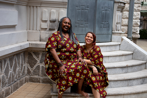 As contadoras de histórias Claudia Marques e Pâmela Cristina sorriem sentadas nos degraus de uma escadaria de pedra diante de um edifício histórico. As duas vestem roupas com a mesma estampa em tons de vinho, amarelo e azul, criando uma identidade visual marcante. O ambiente de arquitetura clássica reforça o clima acolhedor da fotografia.