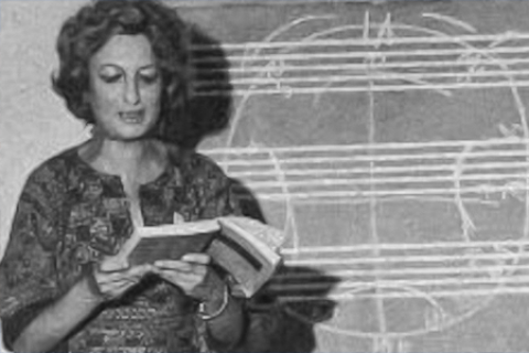 Fotografia em preto e branco da compositora e professora Esther Scliar. Ela aparece lendo um livro aberto, diante de um quadro com diversas linhas e formas geométricas desenhadas a giz. A sombra de seu perfil é projetada sobre o quadro, ao lado das anotações.