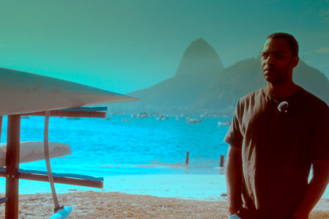 Still do filme Racismo. Um homem aparece em primeiro plano, à direita, usando camiseta escura e olhando para fora do quadro. Ao fundo, vê-se a Baía de Guanabara, com o Pão de Açúcar em destaque entre os morros. À esquerda, parte de uma estrutura com equipamentos náuticos ocupa a lateral da imagem. A cena tem predominância de tons azulados e esverdeados.