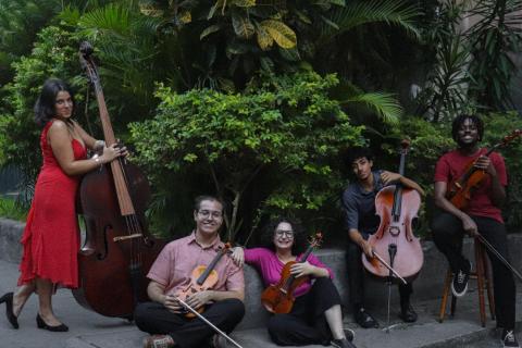 Integrantes do Coletivo Ipê posam com seus instrumentos em uma área externa cercada por vegetação. Os cinco músicos aparecem com instrumentos de cordas e arco de diferentes tamanhos, entre eles contrabaixo e violoncelo. Alguns estão sentados no chão e outros em pé ou sobre um banco, tendo árvores e plantas ao fundo.