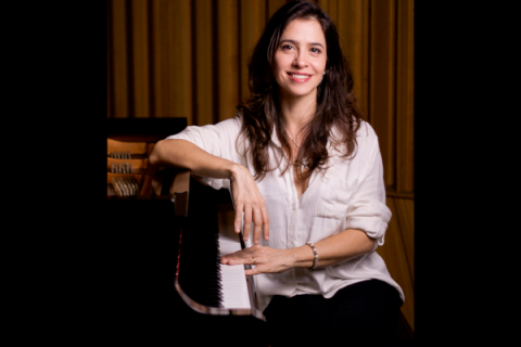 A pianista Marina Spoladore sorri para a câmera enquanto está sentada ao lado de um piano de cauda. Veste camisa branca de mangas dobradas e apoia um dos braços sobre o instrumento, mantendo a outra mão próxima às teclas. Ao fundo, painéis de madeira em tons quentes compõem o ambiente, reforçando a atmosfera acolhedora do retrato.
