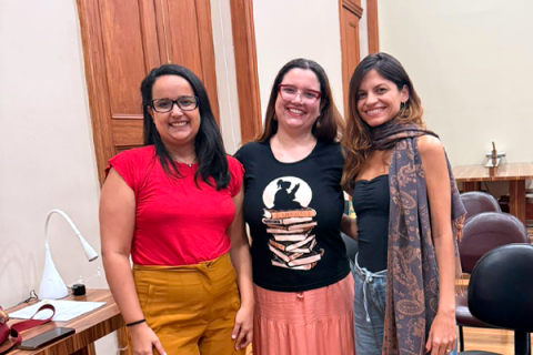 As escritoras e mediadoras Beatriz Ras, Carolina Pessoa e Nicole Ayres posam juntas, sorrindo para a câmera, em uma sala do CCJF. As três aparecem lado a lado, em pé, diante de portas de madeira, vestindo roupas em tons de vermelho, preto, rosa e cinza. Ao fundo, mesas e cadeiras compõem o ambiente.