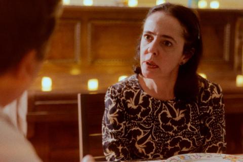 Still do filme O que resta de nós?. A atriz Sílvia Buarque aparece sentada à mesa, voltada para uma pessoa desfocada em primeiro plano. Com expressão atenta e séria, olha diretamente para o interlocutor enquanto parece conversar. Veste uma blusa estampada em preto e branco. Ao fundo, um ambiente iluminado por pequenas luzes em tons quentes reforça a atmosfera intimista da cena.