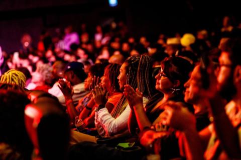 Público durante uma sessão do Encontro de Cinema Negro Zózimo Bulbul. Em primeiro plano, pessoas sentadas lado a lado aplaudem, voltadas para o palco ou a tela fora da imagem. Ao fundo, uma plateia numerosa ocupa o espaço, sob iluminação predominantemente vermelha e alaranjada.