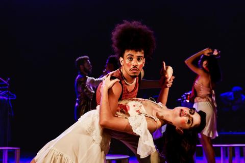 Cena do espetáculo teatral Um Homem Célebre. Em primeiro plano, um ator de cabelos volumosos, usando blusa vermelha sem mangas, sustenta uma atriz em um passo de dança, inclinando-a para trás. Ela veste um figurino claro, com detalhes florais e rendas. O ator olha diretamente para a câmera com expressão de surpresa. Ao fundo, outros artistas aparecem em movimento, sob iluminação cênica em tons de roxo e azul.
