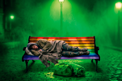 Em uma cena noturna envolta por névoa verde, uma pessoa com roupas desgastadas dorme encolhida sobre um banco pintado com as cores do arco-íris. No chão de pedras, à frente do banco, estão uma mochila e um cobertor enrolado. Ao fundo, postes de iluminação atravessam a névoa.
