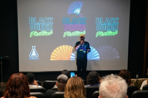 Foto do Black Queer Festival. Em uma sala de cinema, uma pessoa de boné e camiseta preta fala ao microfone diante de uma plateia sentada. Ao fundo, uma grande tela exibe três vezes o logotipo colorido “Black Queer Festival”, acompanhado de ilustrações de leques.