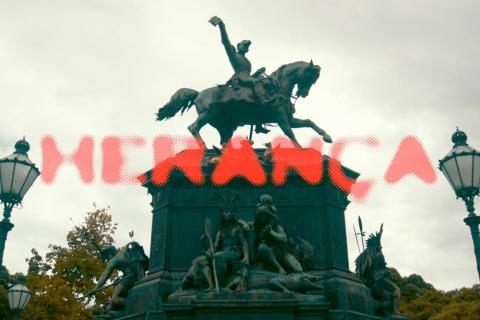 Cartaz do curta-metragem Herança. Fotografia de um monumento equestre, com a estátua de um homem montado a cavalo, erguendo um dos braços. Na base do monumento, aparecem esculturas de figuras humanas sentadas. Sobre a imagem, o título “Herança” está escrito em letras grandes e vermelhas, com efeito pontilhado. Ao fundo, céu nublado e árvores.