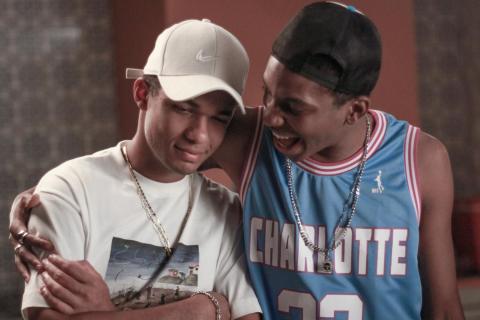Cena do filme Nosso Sonho. À esquerda, o ator Juan Paiva, no papel de Buchecha, usa boné branco, camiseta clara e corrente dourada. Ao seu lado, Lucas Penteado, que interpreta Claudinho, veste uma regata azul de basquete e boné preto virado para trás. Lucas abraça Juan pelos ombros e sorri enquanto fala com ele, que mantém o olhar abaixado e um sorriso discreto.